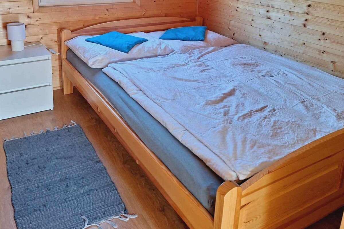 A single bed with a wooden frame and a grey mattress, accompanied by a grey rug and a white bedside table with a lamp.
