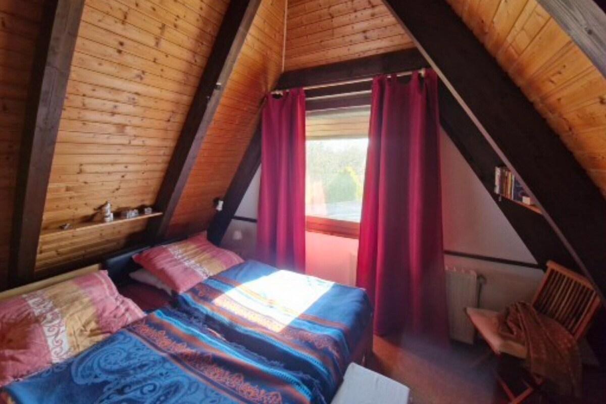 Una habitación con techo inclinado, vigas de madera, cama con ropa de cama estampada, una silla y una ventana con cortinas.