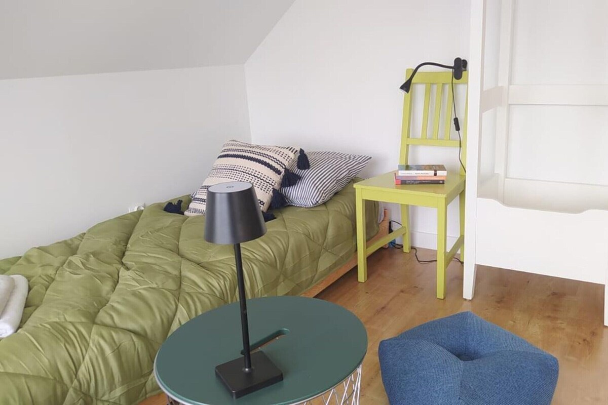 A small, well-lit room with a bed, a table, a chair, and a blue ottoman.