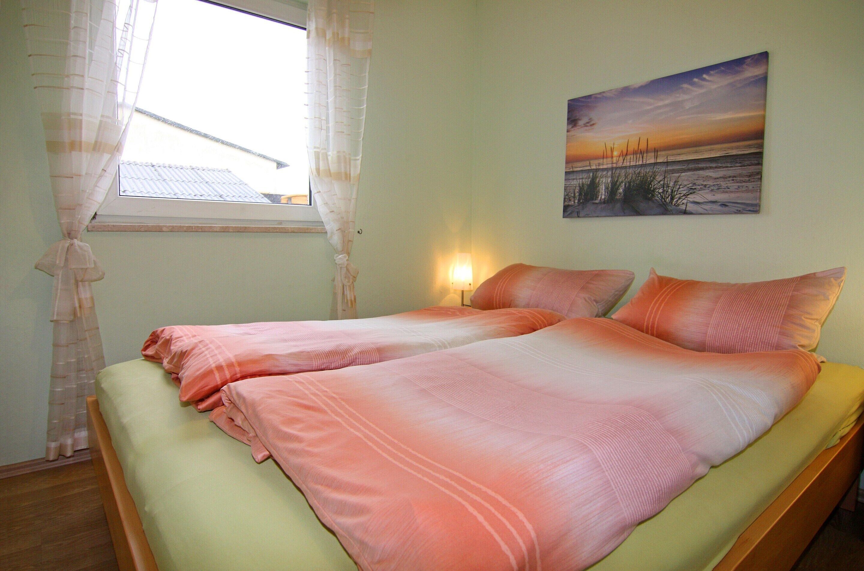 Ein Schlafzimmer mit Bett, einem Vorhangfenster, einer Wandlampe und einem Gemälde mit Sonnenuntergang am Strand.