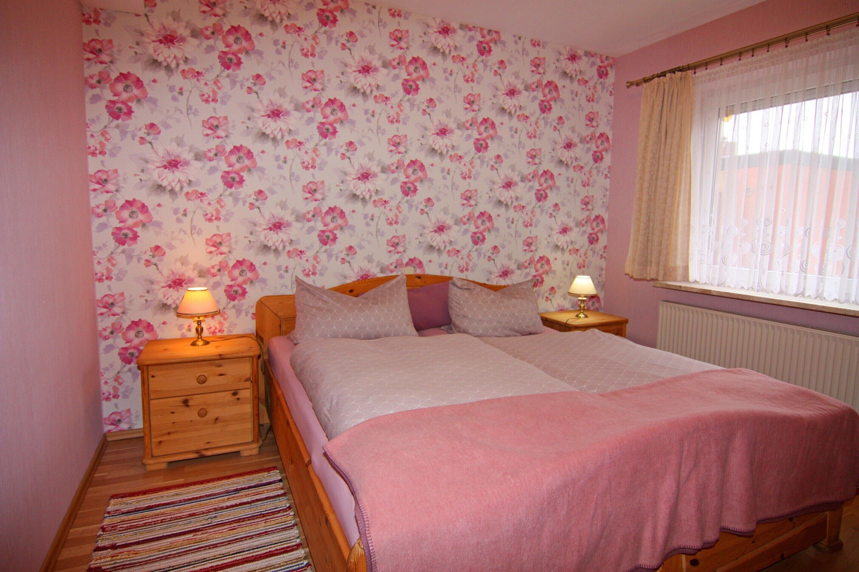 A bedroom with a floral wallpaper, a bed with a pink blanket, two bedside tables with lamps, and a window with curtains.