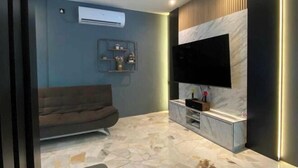 A modern living room with a flat-screen TV mounted on a marble wall, a sofa, and a small shelf with decorative items. - 4BR 16pax LYL Jaccuzi Private Pool House ipoh (Ipoh)