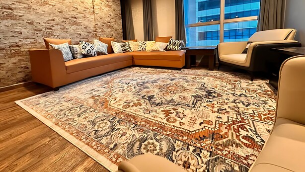 A modern living room with a sectional sofa, a patterned rug, a leather armchair, and a large window with a view of a building. - Centaurus Ultra Luxury Apartments (Islamabad)
