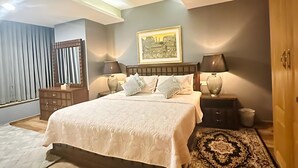 A bedroom with a bed, bedside lamps, a nightstand, a rug, a dresser, and a painting on the wall. - Centaurus Ultra Luxury Apartments (Islamabad)