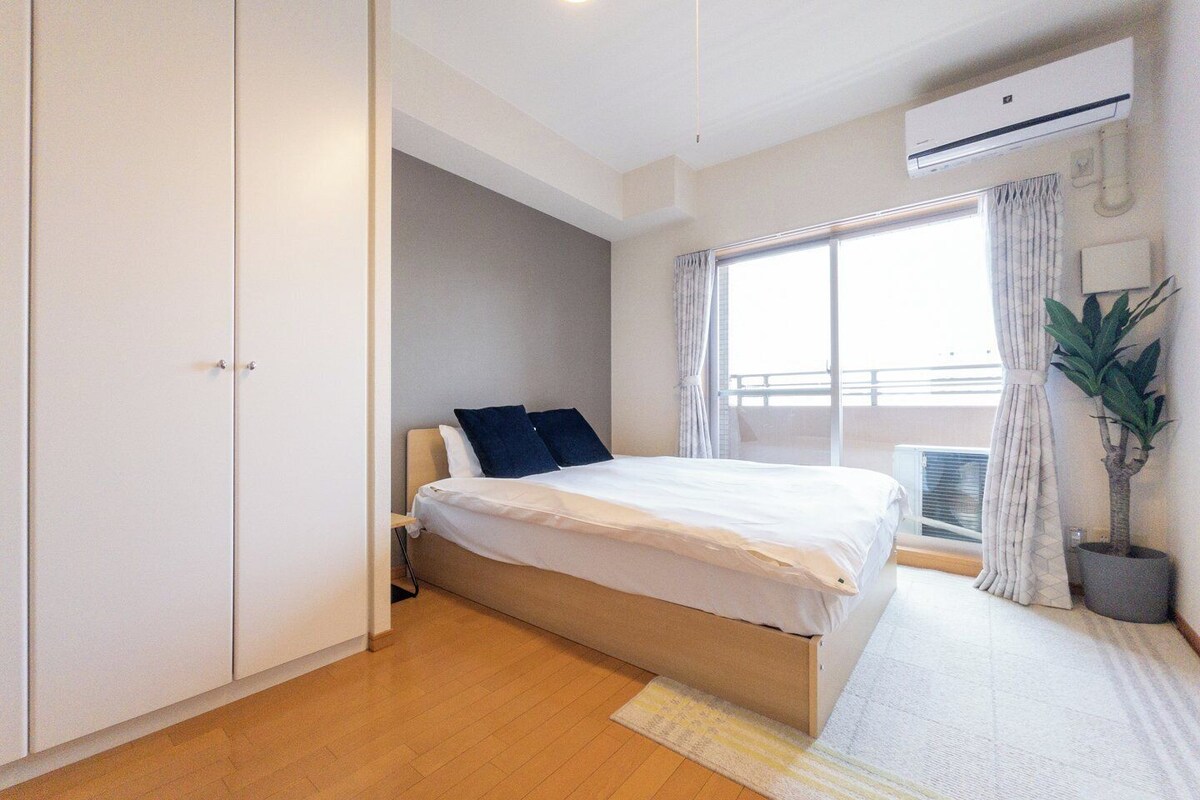 A bedroom with a bed, wardrobe, window, air conditioning unit, and a potted plant.
