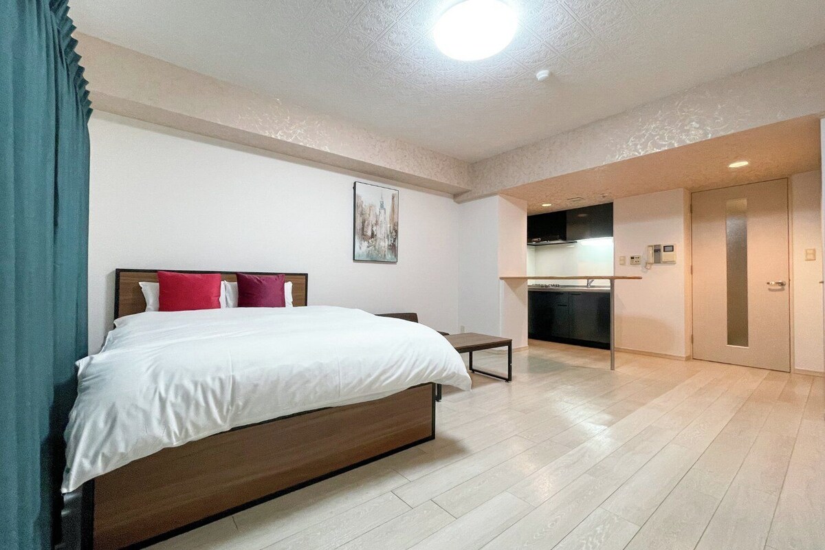 A hotel room with a bed, a small table, a kitchenette, and a door.
