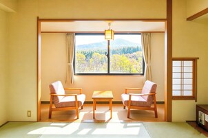 Una habitación japonesa tradicional con suelo de tatami, una mesa baja, dos sillas, un ventanal con vistas a las montañas y una puerta corredera.
