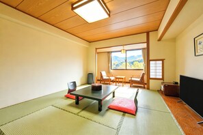Una habitación japonesa tradicional con suelo de tatami, una mesa baja, sillas, un televisor y vistas a montañas a través de un ventanal grande.
