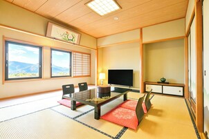 Una habitación japonesa tradicional con suelo de tatami, una mesa baja, sillas, un televisor y vistas a las montañas a través de amplias ventanas.