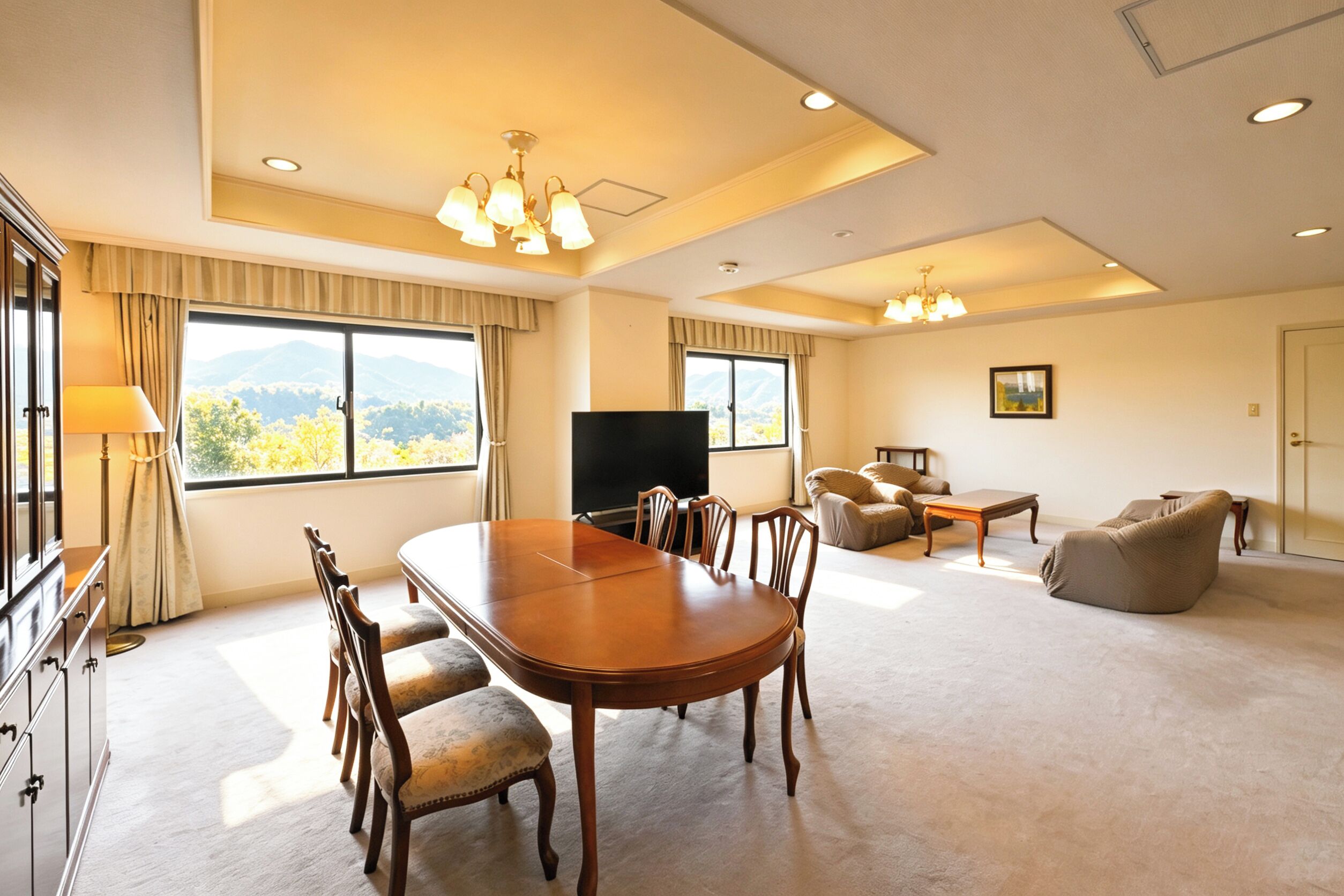 A spacious living area with a large wooden dining table, comfortable seating, a flat-screen TV, and a view of mountains through large windows.