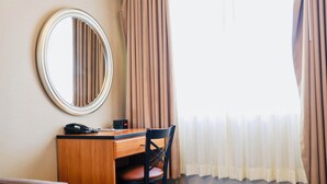 Quarto de hotel com secretária de madeira, uma cadeira, um espelho redondo, telefone e janela com cortinas.