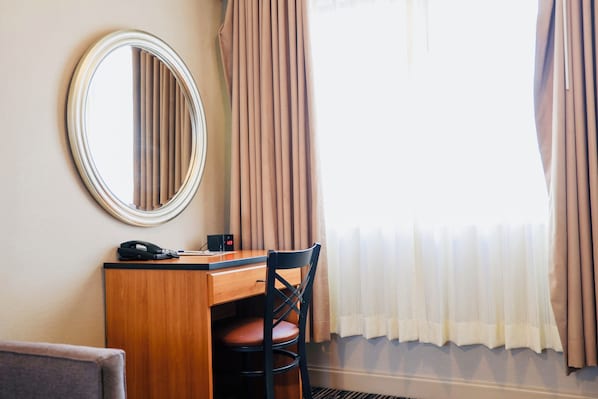 A hotel room with a wooden desk, a chair, a round mirror, a telephone, and a window with curtains.