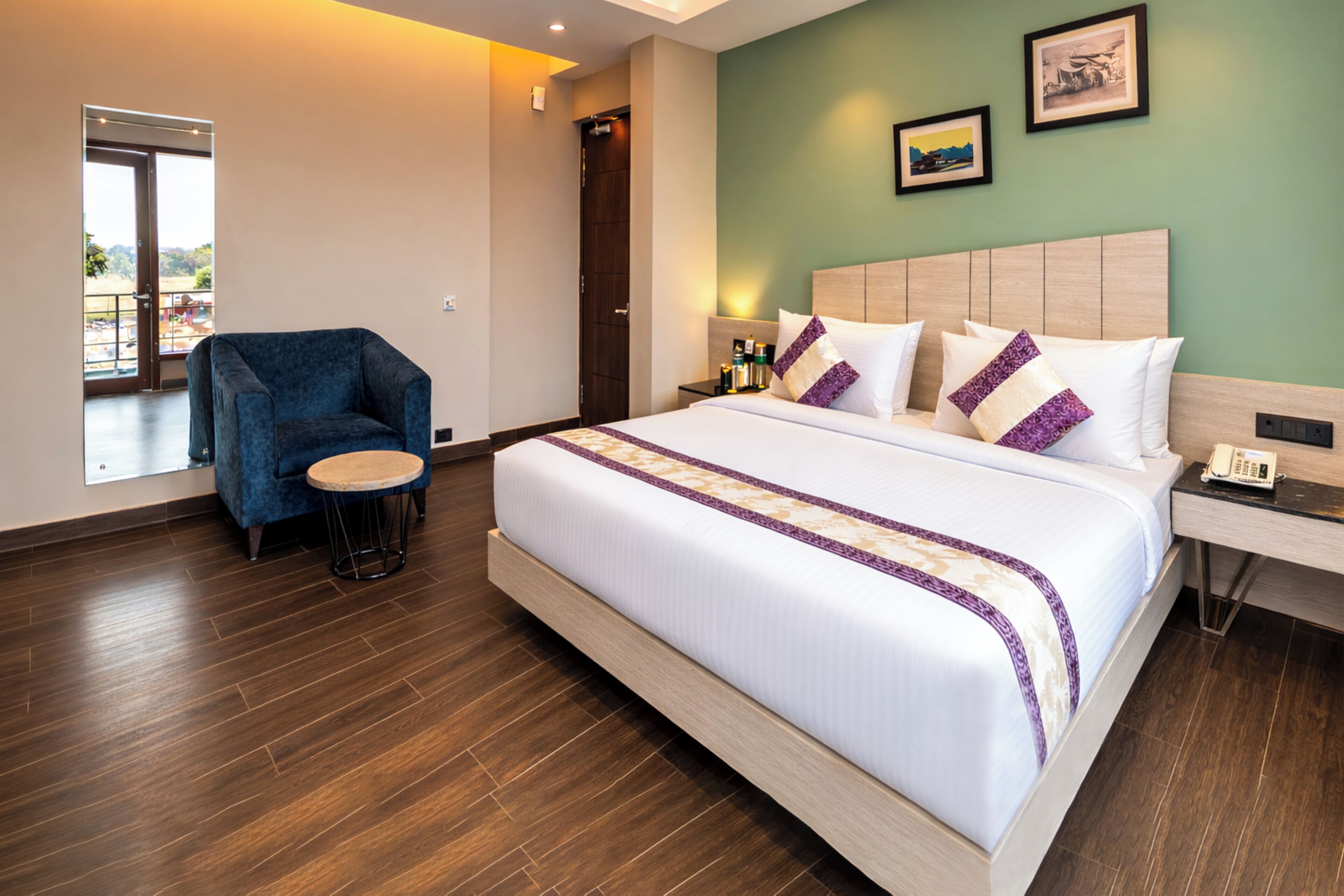 A hotel room with a large bed, a blue armchair, a small round table, a nightstand with a phone, and a door leading to a balcony.