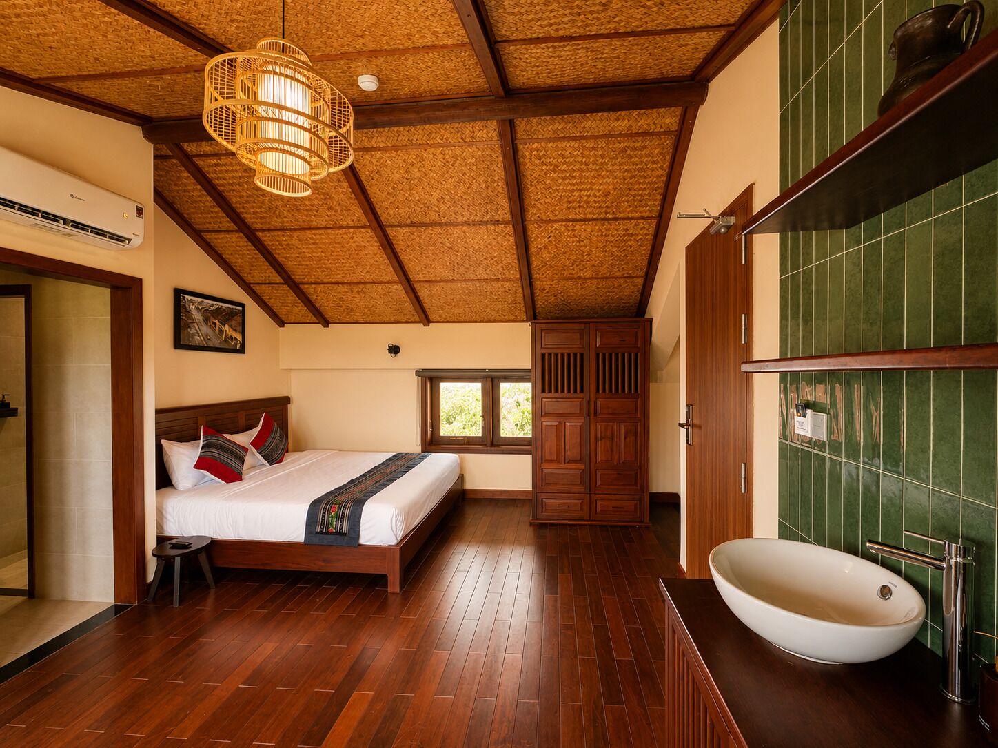 Una habitación con cama, suelo de madera, un ventanal grande y baño con lavamanos y espejo.