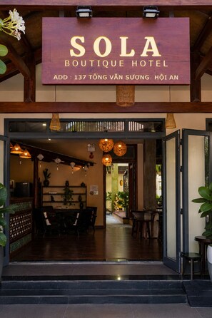 La entrada del Hotel Boutique Sola, con fachada de madera, un letrero y un área de recepción con mesas y sillas.