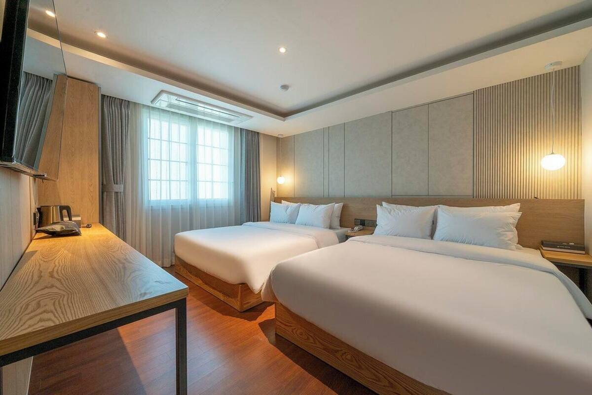 A hotel room with two beds, a wooden desk, a TV, and a window with curtains.