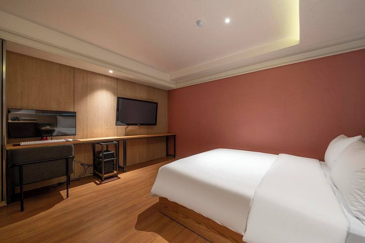 A hotel room with a bed, a desk with a computer, a TV, and a wooden floor.