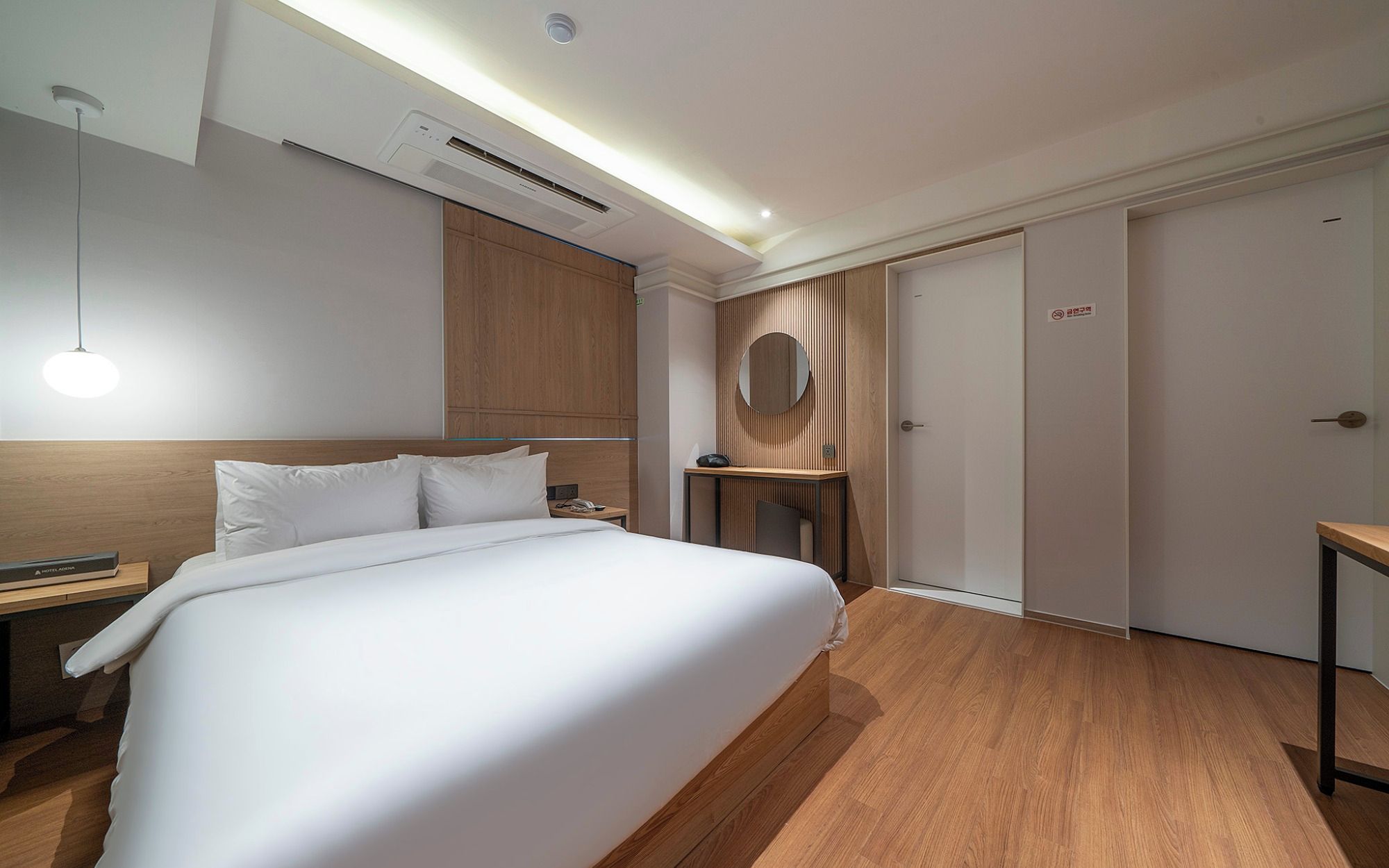 A hotel room with a large bed, a bedside table, a desk with a chair, a round mirror, and a wardrobe.