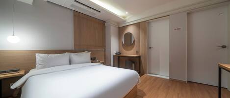 A hotel room with a large bed, a bedside table, a desk with a chair, a round mirror, and a wardrobe.