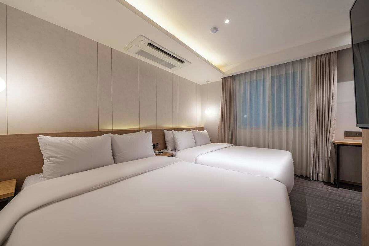 A hotel room with two beds, a desk, a TV, and an air conditioning unit.