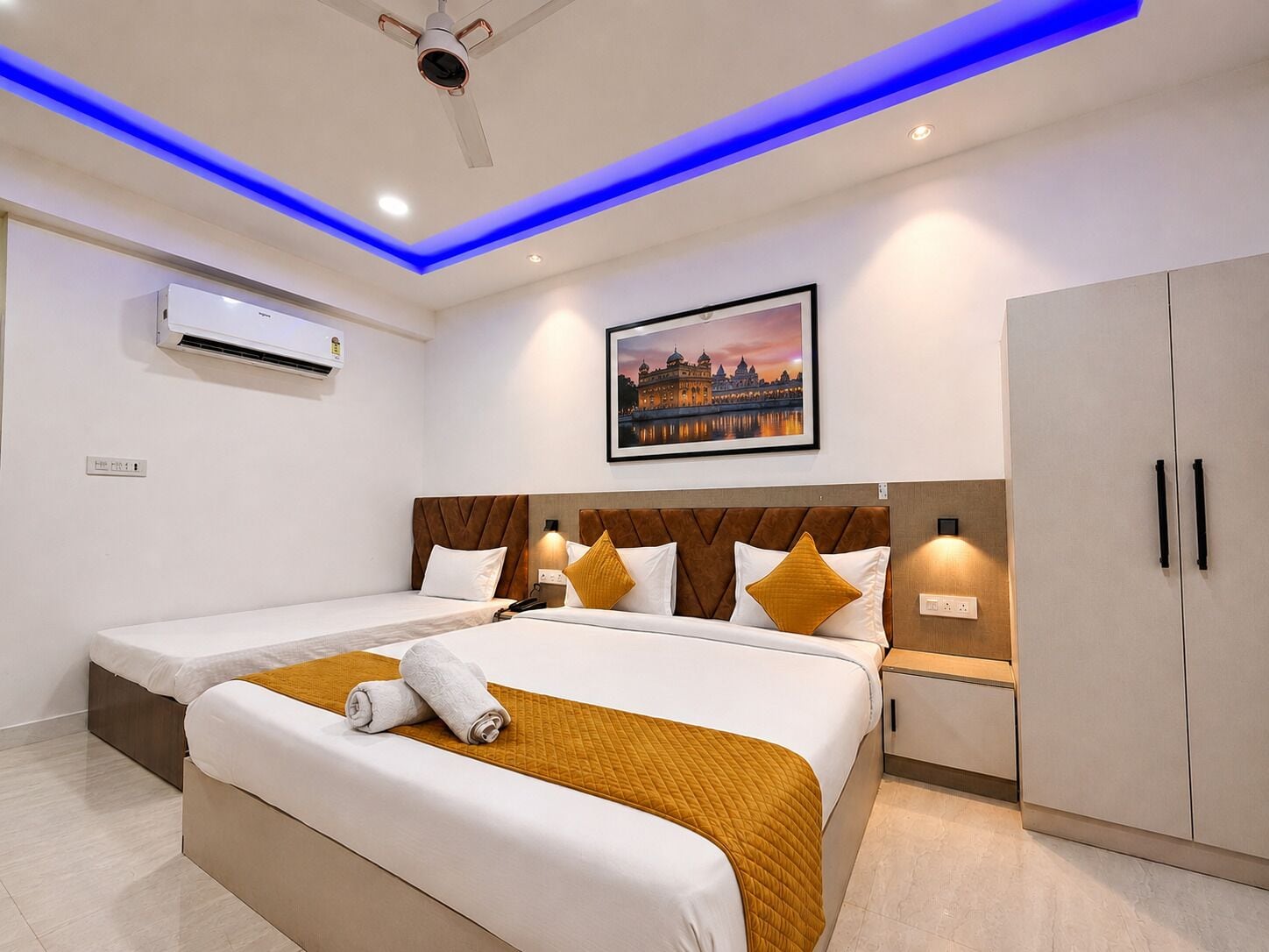 A hotel room with two beds, a ceiling fan, an air conditioning unit, a framed picture of a building, and a wardrobe.
