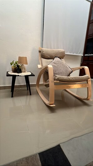 Ein Schaukelstuhl mit Kissen, ein kleiner Tisch mit Lampe und Blumenvase sowie eine schlichte Wand.