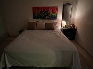 Ein Bett mit weißer Bettdecke und zwei Kissen, ein Bild an der Wand, ein Nachttisch mit Lampe und ein Spiegel.