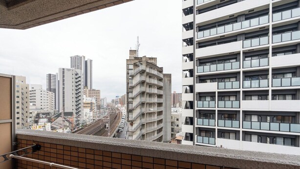 A high-rise building with multiple balconies, a tiled balcony floor, and a view of a cityscape with other buildings and a railway track. - AXIA (Osaka)