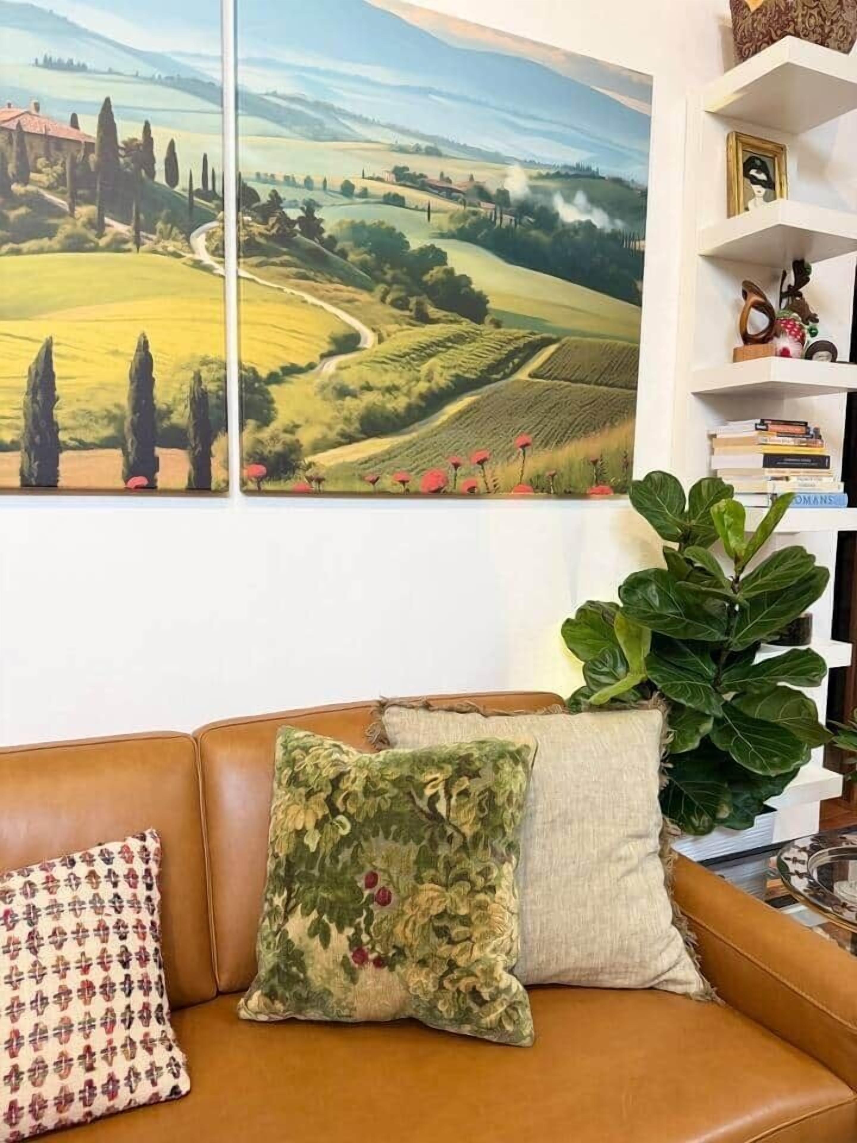 A living room with a brown leather sofa, two patterned cushions, a large landscape painting, a bookshelf with books, and a large green plant.