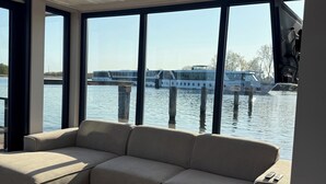 A modern interior with a large window overlooking a water body, a beige sofa, and two round side tables. - Unique stay on the water, houseboat Loosdrecht (Loosdrecht)