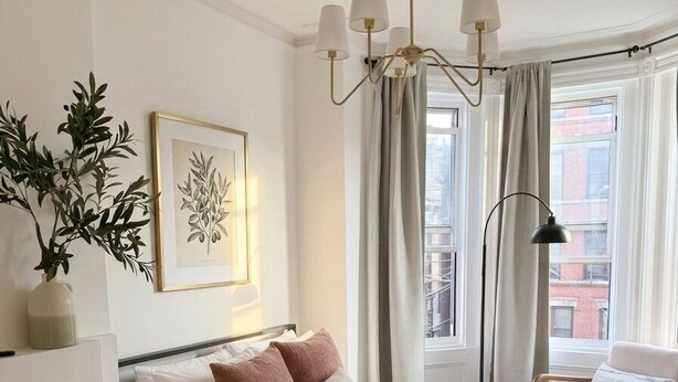 Ein Hotelzimmer mit Bett, Nachttisch, Lampe, Fenster mit Vorhängen und einer Pflanze.
