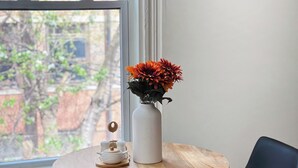 Ein runder Holztisch mit einer weißen Vase und orangefarbenen Blumen, zwei schwarze Stühle und ein Fenster mit Blick auf Gebäude und Bäume.
