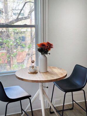 Ein runder Holztisch mit einer weißen Vase und orangefarbenen Blumen, zwei schwarze Stühle und ein Fenster mit Blick auf Gebäude und Bäume.