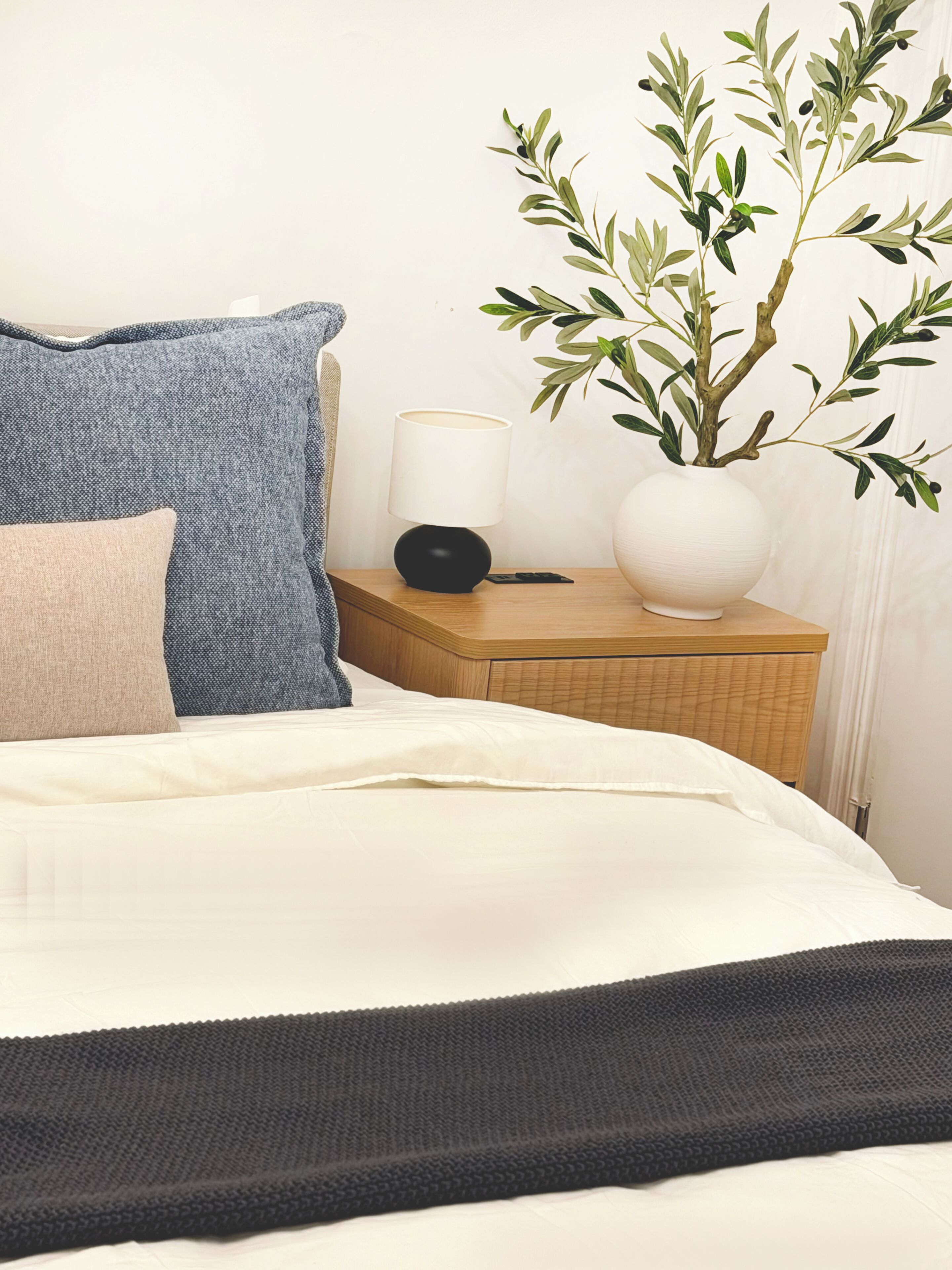 En pænt redt seng med et hvidt og sort tæppe, en blå pude, en beige pude, en hvid vase med grønne blade, og en sort lampe på et trænatbord.