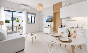 Ein modernes Wohnzimmer mit einer Couch, einem Esstisch für zwei Personen und einer Küchenzeile mit weißen Schränken sowie einem Fernseher.