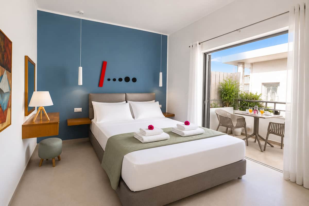 A modern hotel room with a large bed, a balcony with a table and chairs, and a view of greenery.