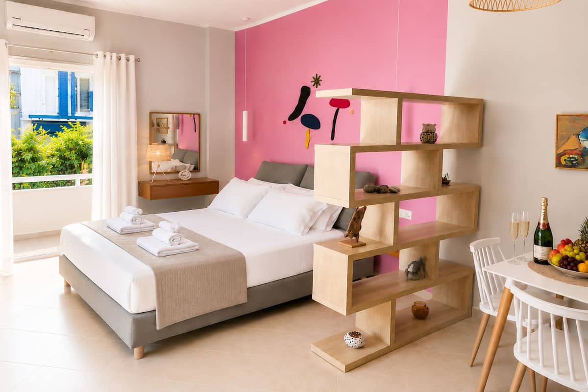 A modern bedroom with a bed, bedside tables, a wooden shelf unit, a dining area with a table and chairs, and a view of greenery outside.