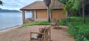 A beachside hut with a thatched roof, wooden chairs, and a view of the water. - Anake Lodge (Cape Maclear)