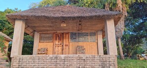 A thatched-roof structure with a wooden door and windows, surrounded by greenery. - Anake Lodge (Cape Maclear)