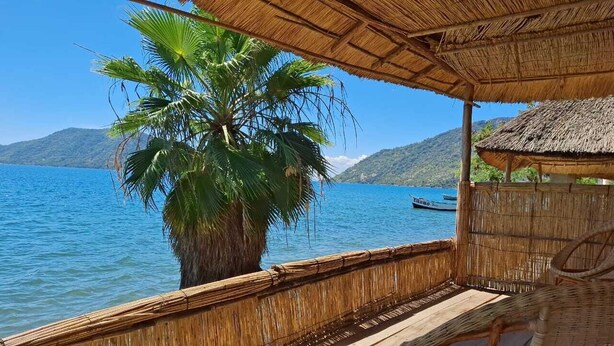 A thatched-roof structure with a view of a calm lake and a distant boat. - Anake Lodge (Cape Maclear)