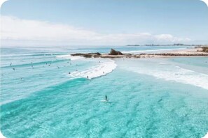 A beach with clear turquoise water, a rocky outcrop, and people swimming and surfing. - The Art Garden: a coastal retreat with sauna and longboard steps from the beach (Tugun)