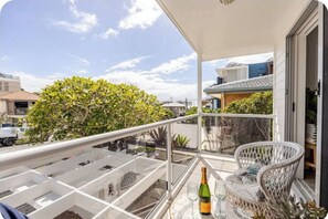 A balcony with a glass railing, a wicker chair, a table with a bottle and glasses, and a view of the street and buildings. - The Art Garden: a coastal retreat with sauna and longboard steps from the beach (Tugun)