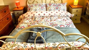 Ein Bett mit floralem Überwurf, ein Metall-Kopfteil, ein Nachttisch aus Holz mit einer Lampe und eine Holzkommode.