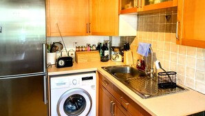 Eine kleine Küche mit Holzschränken, einem Edelstahl-Kühlschrank, einer Waschmaschine und einer gefliesten Spritzschutzwand.