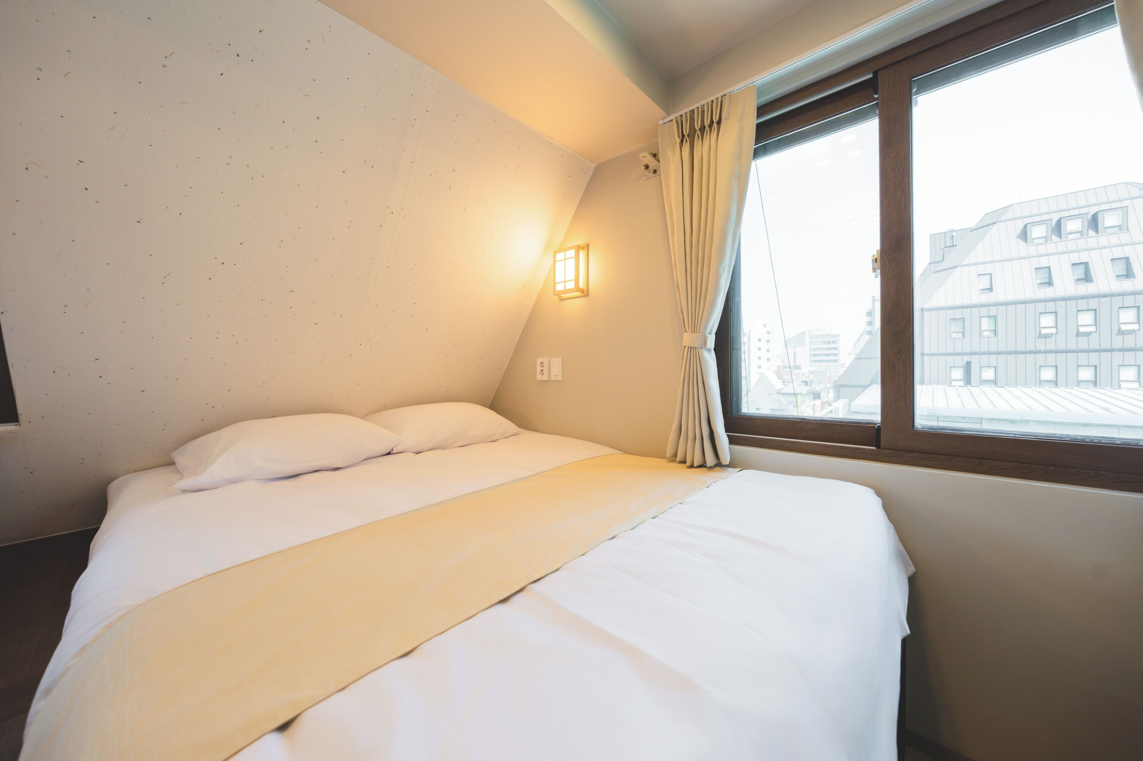 Ein Hotelzimmer mit Bett, einem Vorhangfenster, einer Wandleuchte und Blick auf Gebäude.