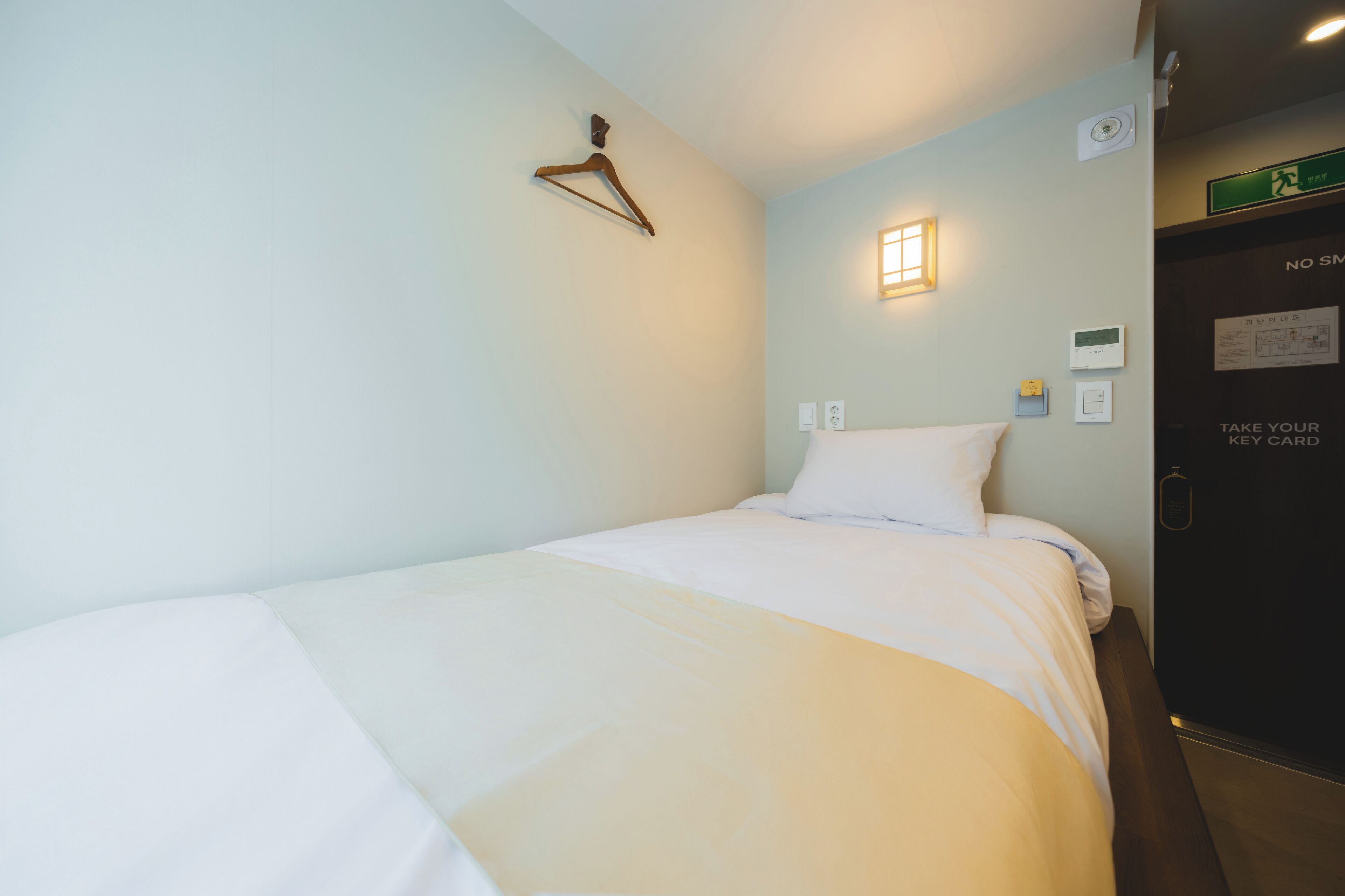 A single bed with white bedding, a wooden hanger on the wall, a small wall-mounted light, and a door with a sign.