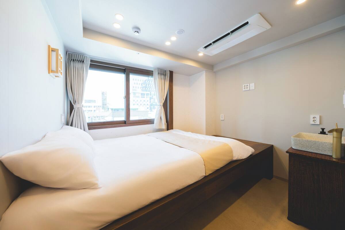 A hotel room with a bed, a window with curtains, an air conditioning unit, and a wooden nightstand with a sink.