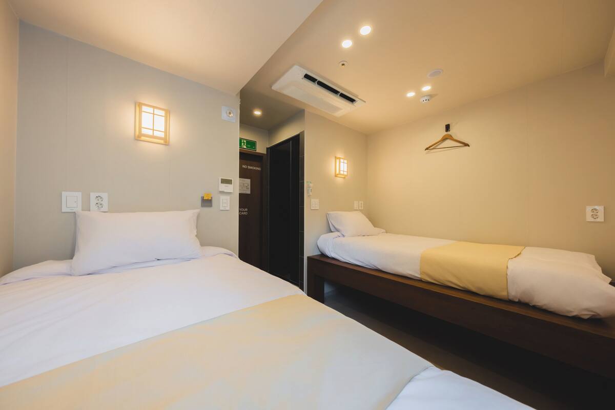 A hotel room with two beds, a wooden bench, a coat hanger, and a door with a 'No Smoking' sign.