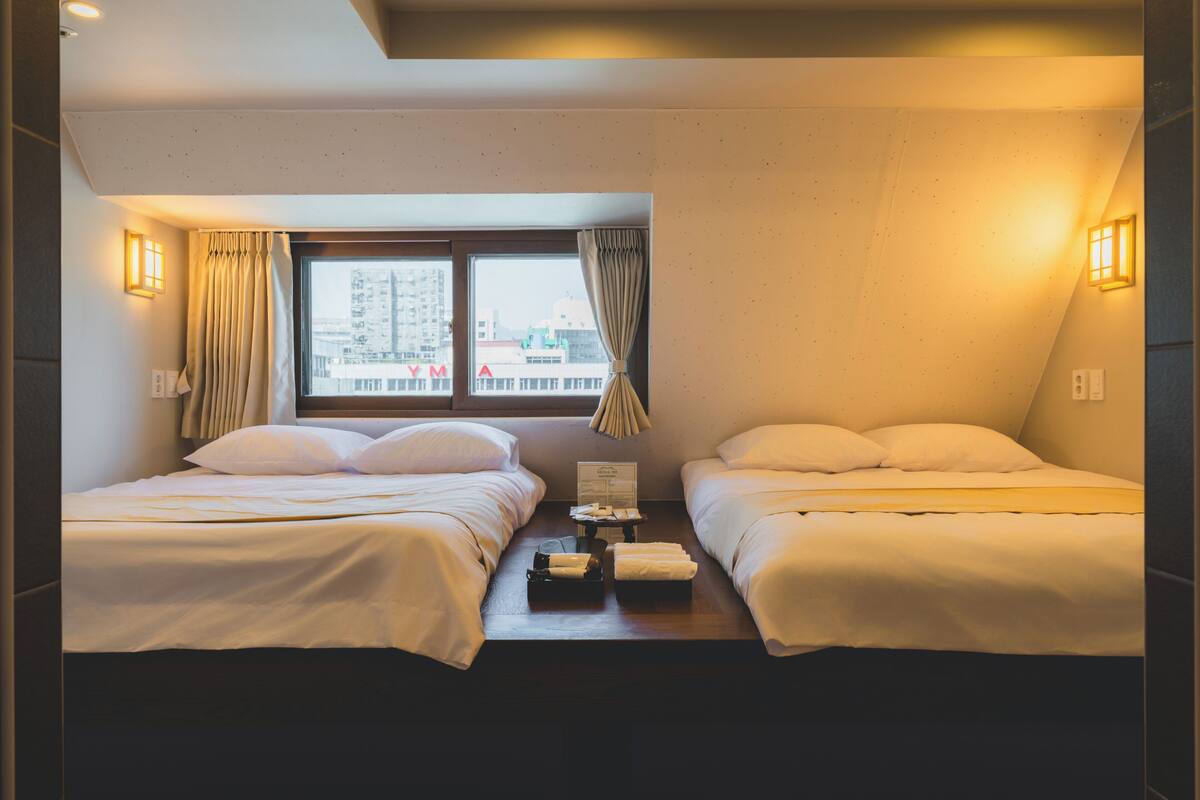 A hotel room with two beds, a window with curtains, a nightstand with a lamp, and a city view.