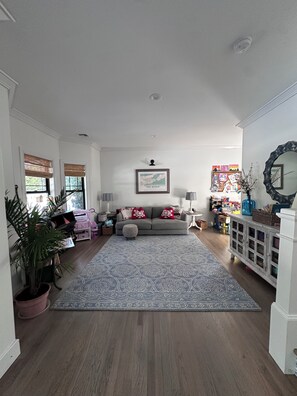 A spacious living room with a grey sofa, patterned rug, and a bookshelf filled with books and decorative items. - East Quogue dream with outdoor turf, sport court and more (East Quogue)